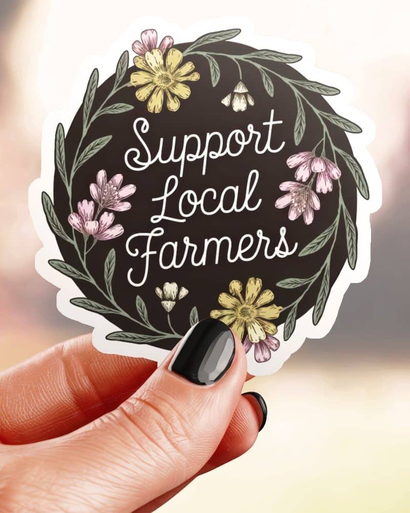 A hand holds a circular sticker that says Support Local Farmers in a floral wreath.