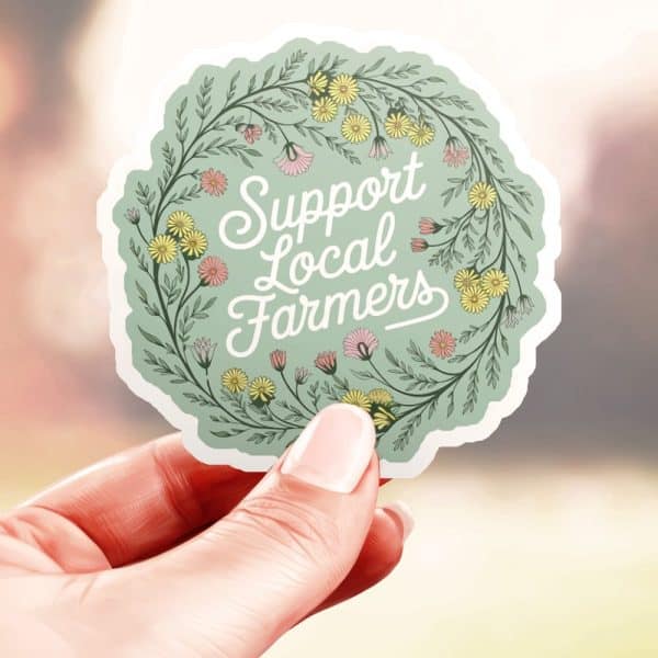 Hand Holding A Round Sticker With A Floral Wreath Design. The Sticker Says Support Local Farmers.