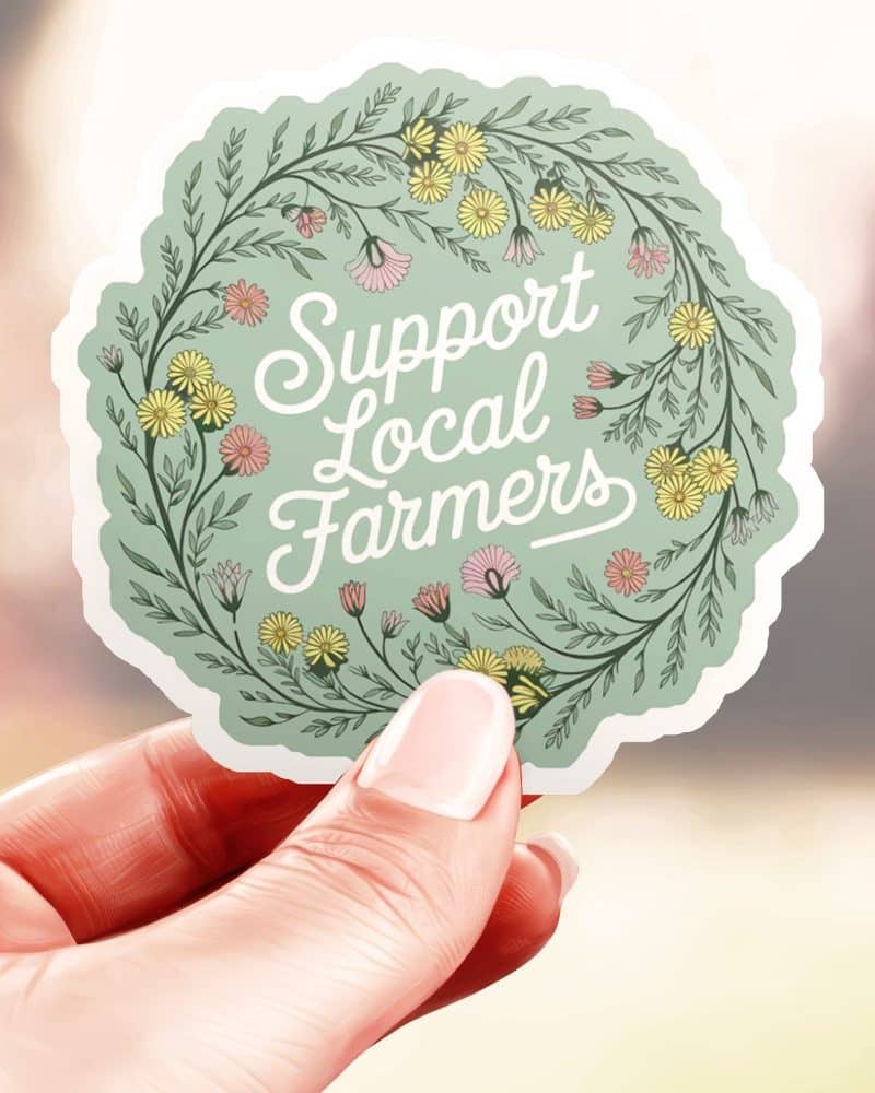 Hand holding a round sticker with a floral wreath design. The sticker says Support Local Farmers.