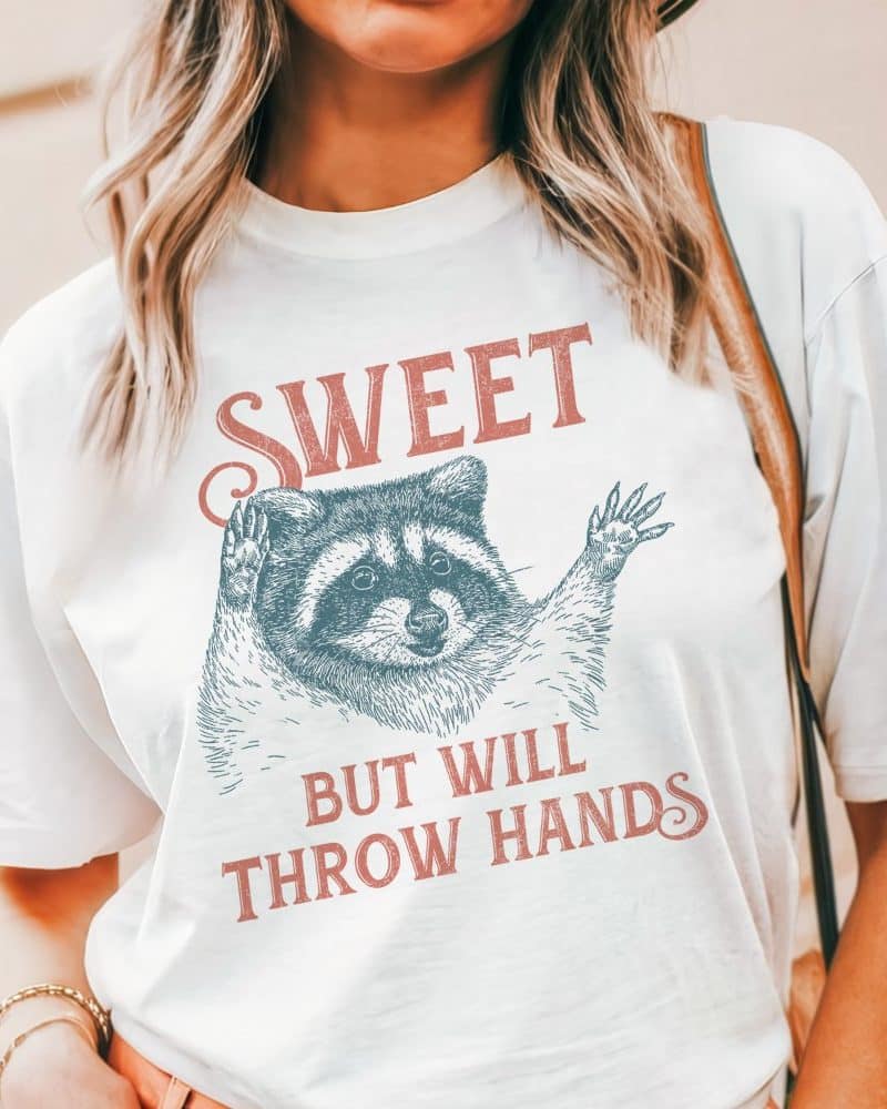 White t-shirt featuring a raccoon graphic and the text Sweet but will throw hands.