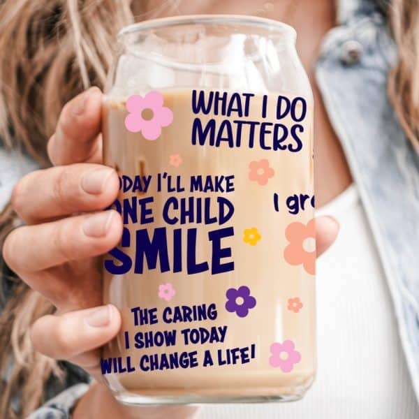 A Hand Holds A Glass Iced Coffee Tumbler With Text That Says, What I Do Matters, And One Child Smile. Smaller Text Below Adds, The Caring I Show Today Will Change A Life. The Tumbler Is Decorated With Small Flowers.