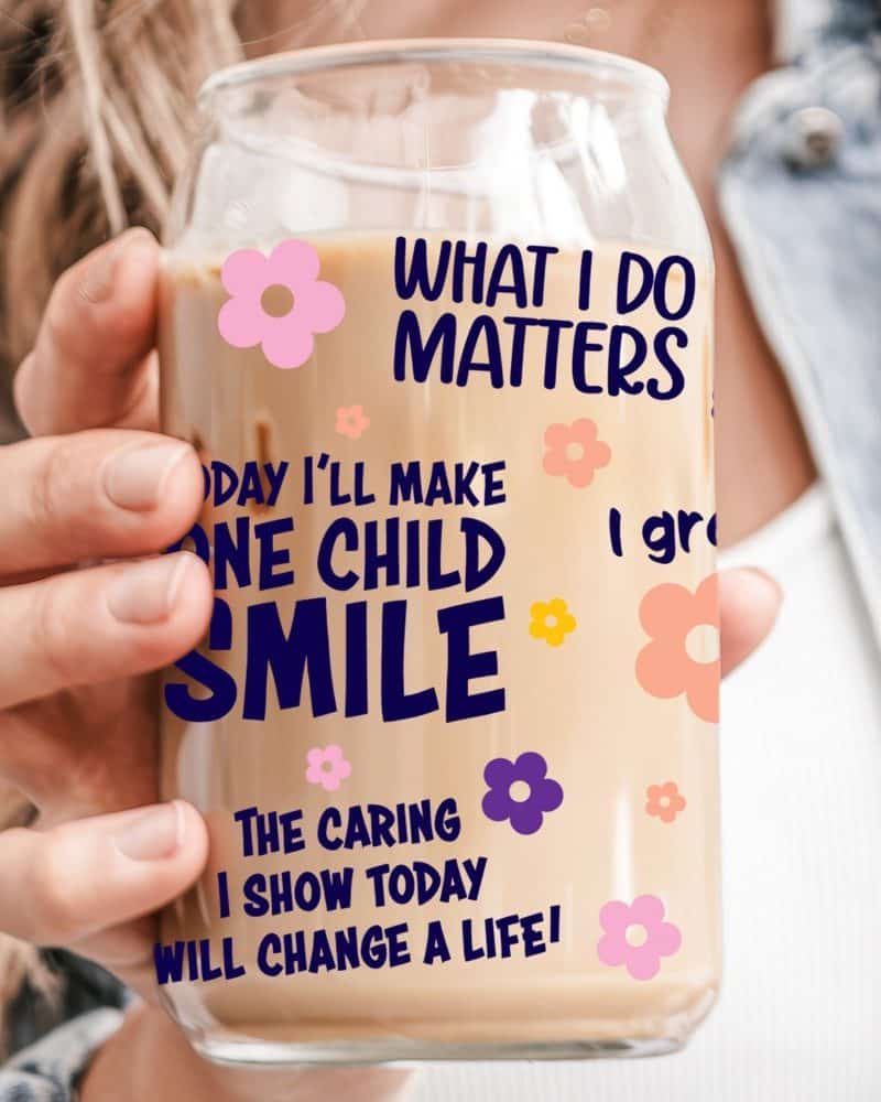 A hand holds a glass iced coffee tumbler with text that says, What I Do Matters, and One Child Smile. Smaller text below adds, The caring I show today will change a life. The tumbler is decorated with small flowers.