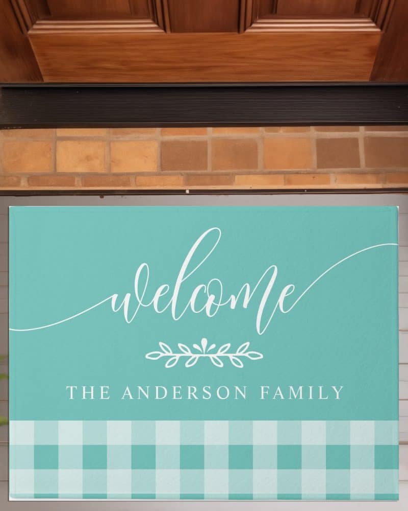 Teal welcome mat with white script lettering that says Welcome and The Anderson Family, and a bottom border of teal and white gingham.