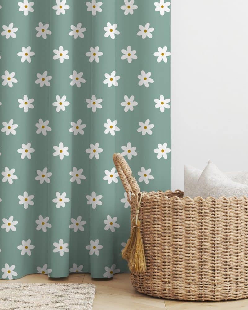 A teal curtain with a white daisy pattern next to a wicker basket holding white pillows.