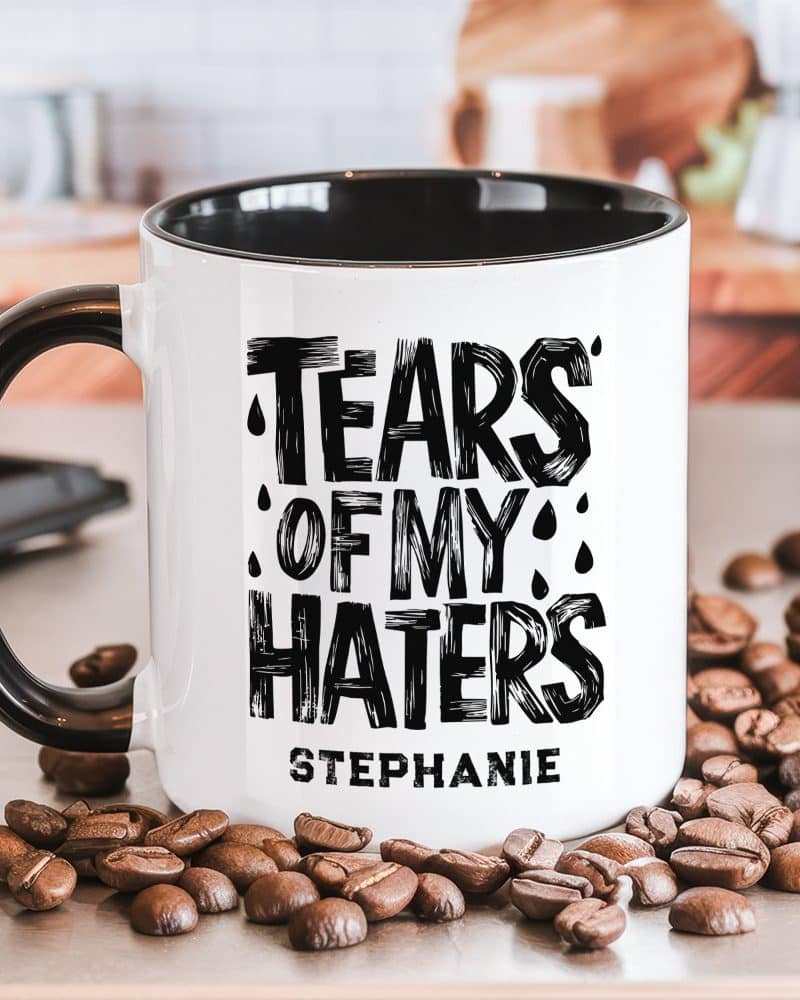 A black and white coffee mug with the text Tears of My Haters and the name Stephanie. Coffee beans are scattered around the mug.
