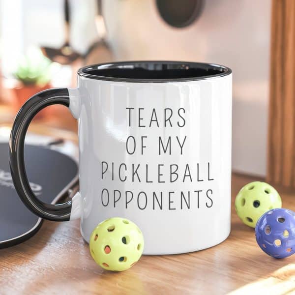 White Coffee Mug With Black Handle And Text Reading Tears Of My Pickleball Opponents, And Three Pickleballs Beside It.