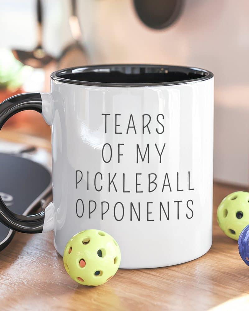 White coffee mug with black handle and text reading Tears of My Pickleball Opponents, and three pickleballs beside it.