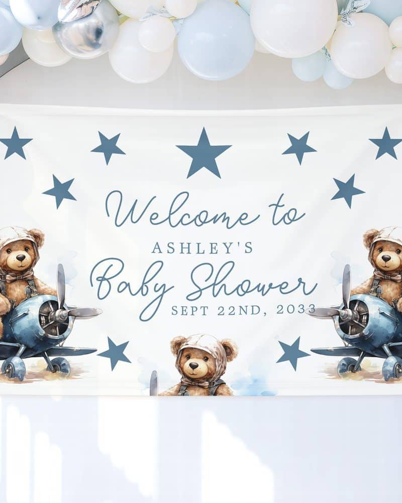 A baby shower welcome sign featuring two teddy bears in aviator hats piloting airplanes, with Ashley's name and the date of the shower.