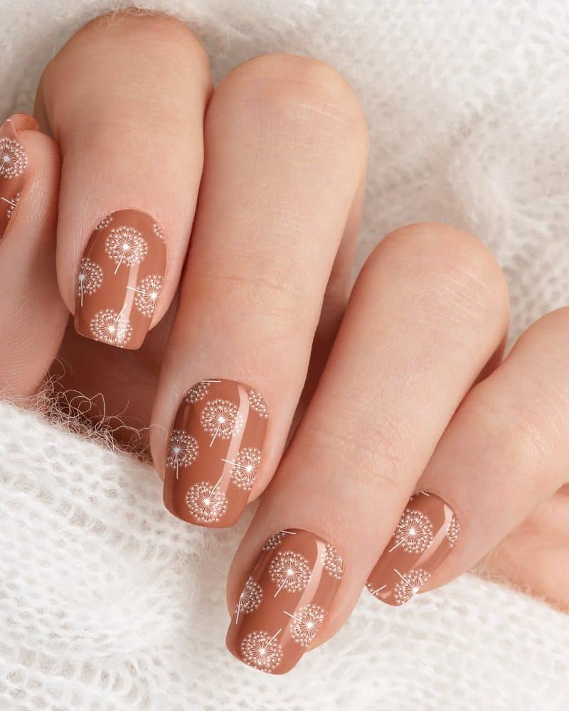 Closeup of a hand with light brown nails decorated with a white dandelion pattern.
