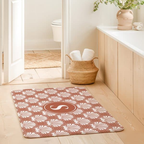 A Terracotta Bath Mat With A White Seashell Pattern And A White Monogrammed Initial In The Center.