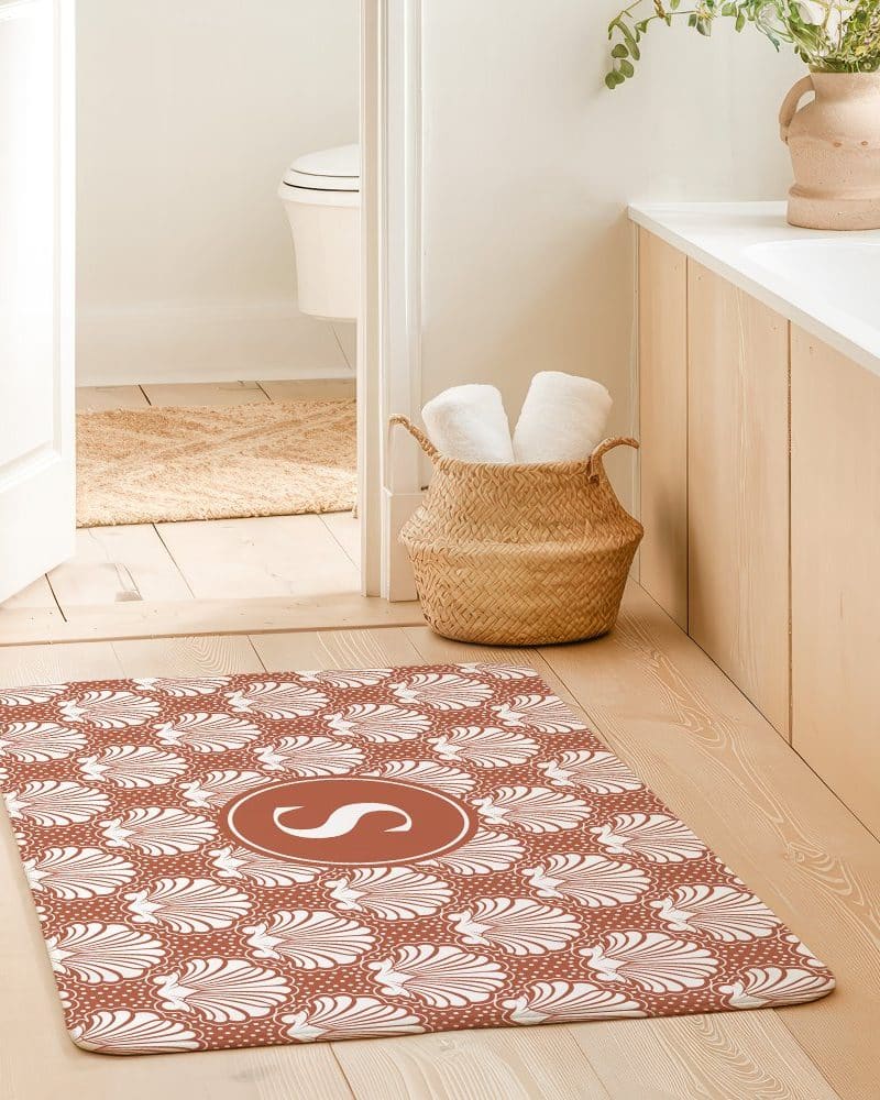 A terracotta bath mat with a white seashell pattern and a white monogrammed initial in the center.