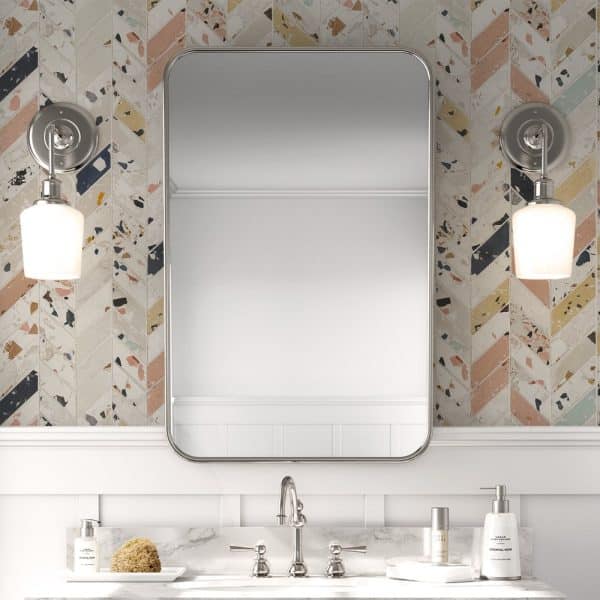 A Rectangular Mirror Hangs Above A Bathroom Sink With A Marble Countertop. The Mirror Is Framed In Brushed Nickel And Is Set Against A Herringbone Patterned Backsplash.