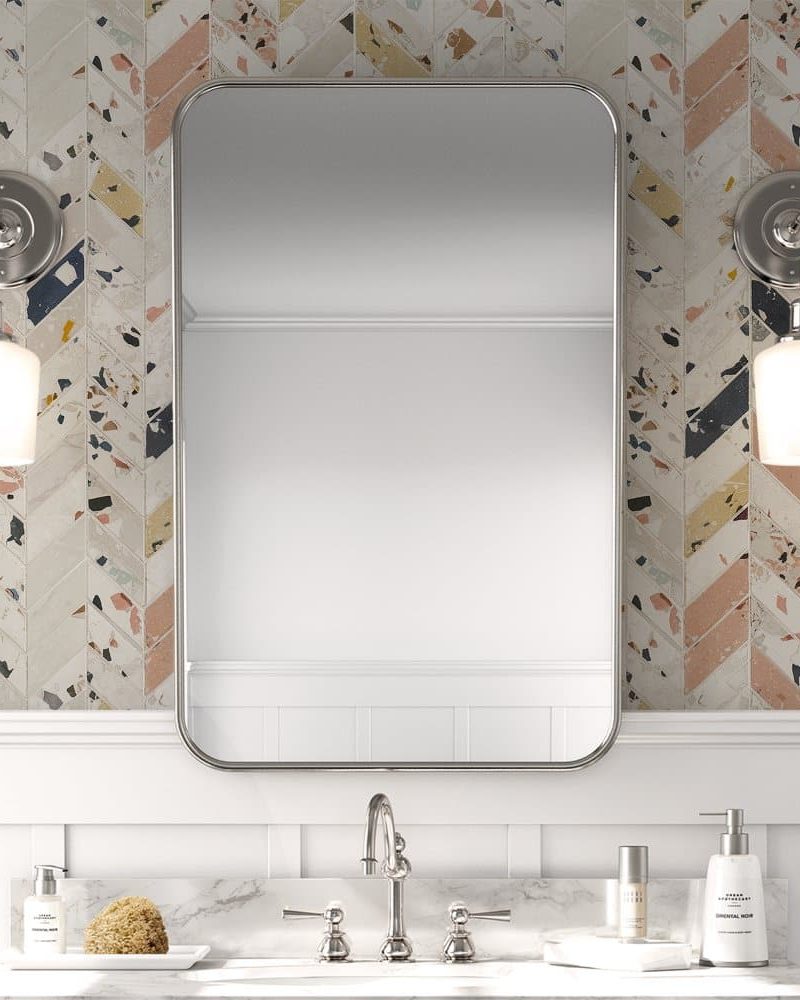 A rectangular mirror hangs above a bathroom sink with a marble countertop. The mirror is framed in brushed nickel and is set against a herringbone patterned backsplash.