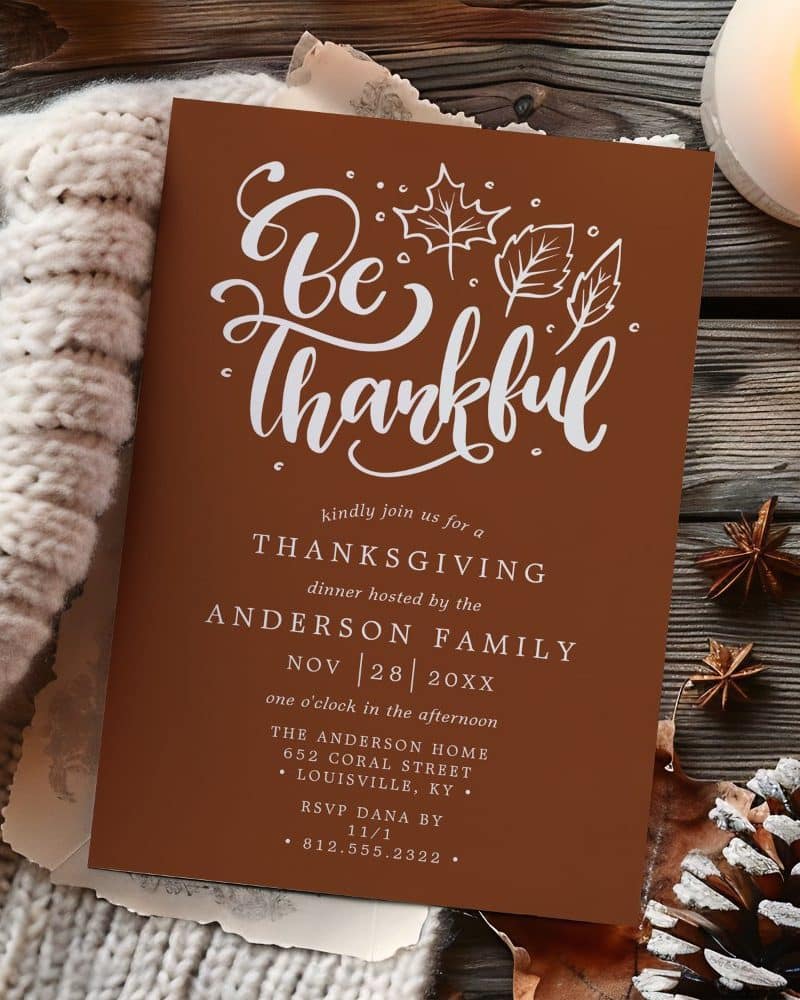 A rust-colored Thanksgiving invitation with white script lettering that says Be Thankful, surrounded by small leaf illustrations.