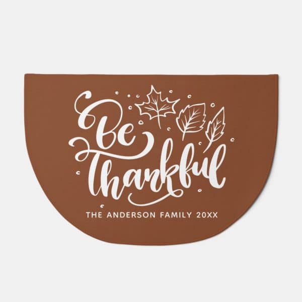 Brown Half-Circle Doormat With White Script Lettering That Says Be Thankful And Small Leaf Illustrations. A Family Name And Year Are Also Printed Below.