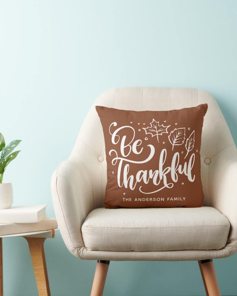 Brown throw pillow with white lettering that says Be Thankful and The Anderson Family. Autumn leaves are also on the pillow.