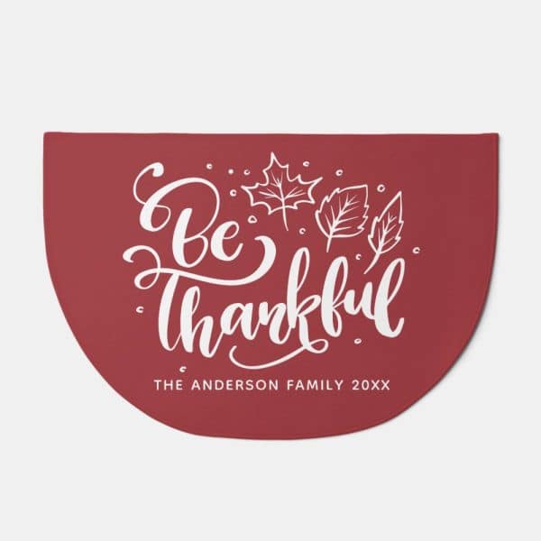 Red Half-Circle Doormat With The Text Be Thankful In A Stylized Script Font And Small Leaf Illustrations.
