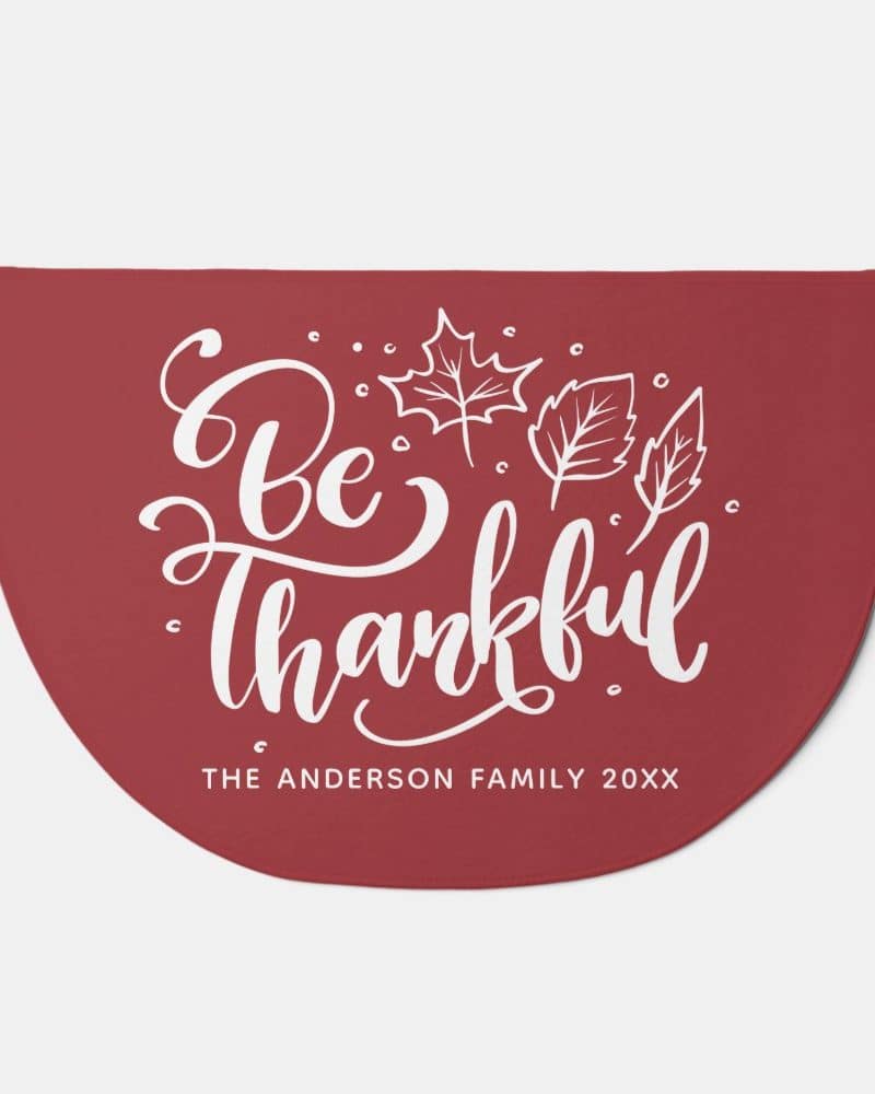 Red half-circle doormat with the text Be Thankful in a stylized script font and small leaf illustrations.