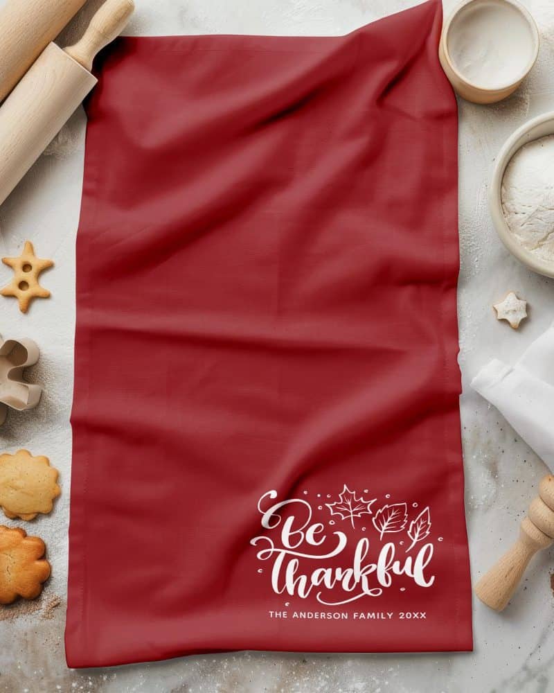 Red kitchen towel with "Be Thankful" in white script and autumn leaves, personalized with a family name and year.