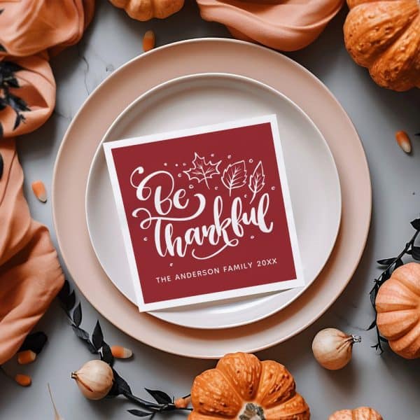 A Maroon Place Card With White Lettering Reading Be Thankful And The Anderson Family.