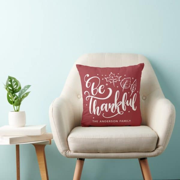 A Maroon Pillow With White Lettering That Says Be Thankful And The Anderson Family Sits On A Beige Armchair.