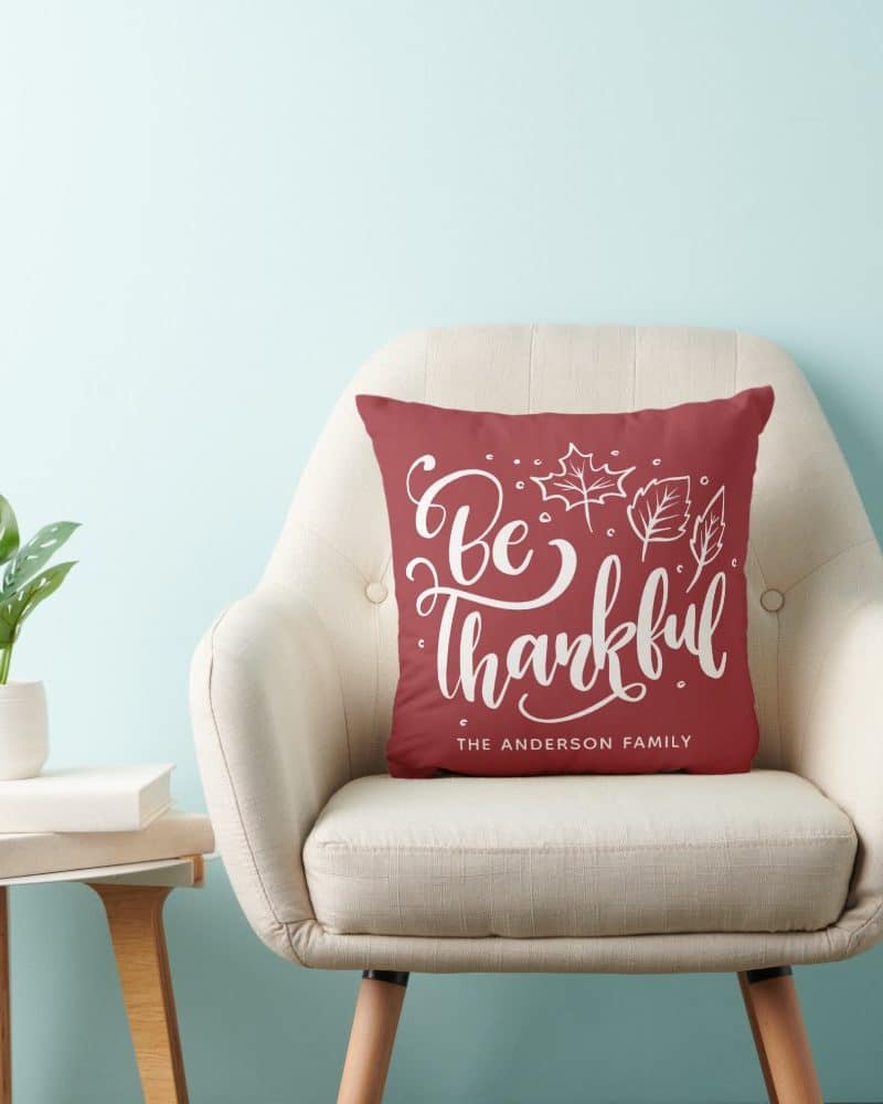 A maroon pillow with white lettering that says Be Thankful and The Anderson Family sits on a beige armchair.