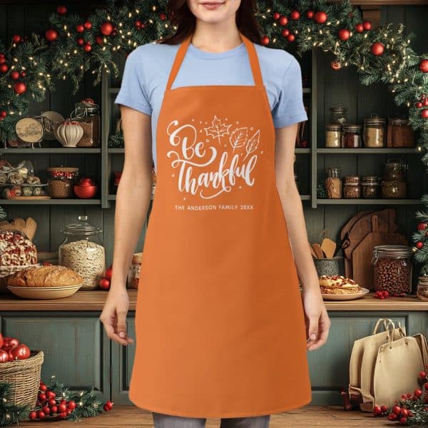 Orange Apron With White Lettering That Says Be Thankful And A Family Name.