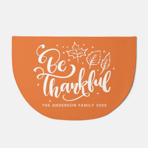 Orange Half-Circle Doormat With The Words Be Thankful In A Stylized Script Font And Small Leaf Illustrations. A Personalized Family Name Is At The Bottom.