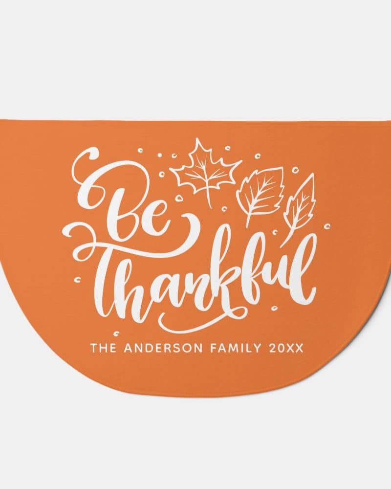 Orange half-circle doormat with the words Be Thankful in a stylized script font and small leaf illustrations. A personalized family name is at the bottom.