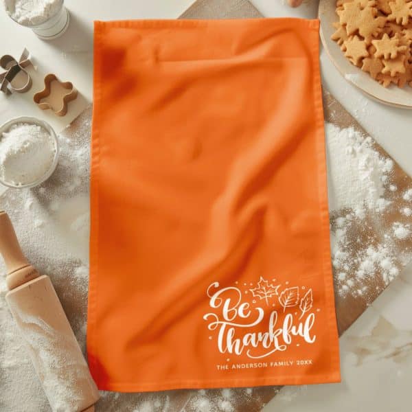 An Orange Kitchen Towel With The Words Be Thankful In White Script Lettering And A Small Autumn Leaf Design. The Anderson Family Name And Year Are Also Printed On The Towel.
