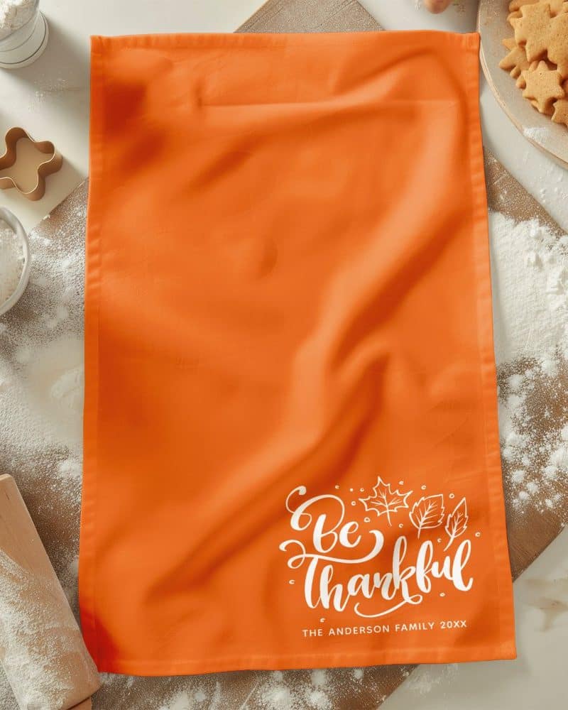An orange kitchen towel with the words Be Thankful in white script lettering and a small autumn leaf design. The Anderson family name and year are also printed on the towel.