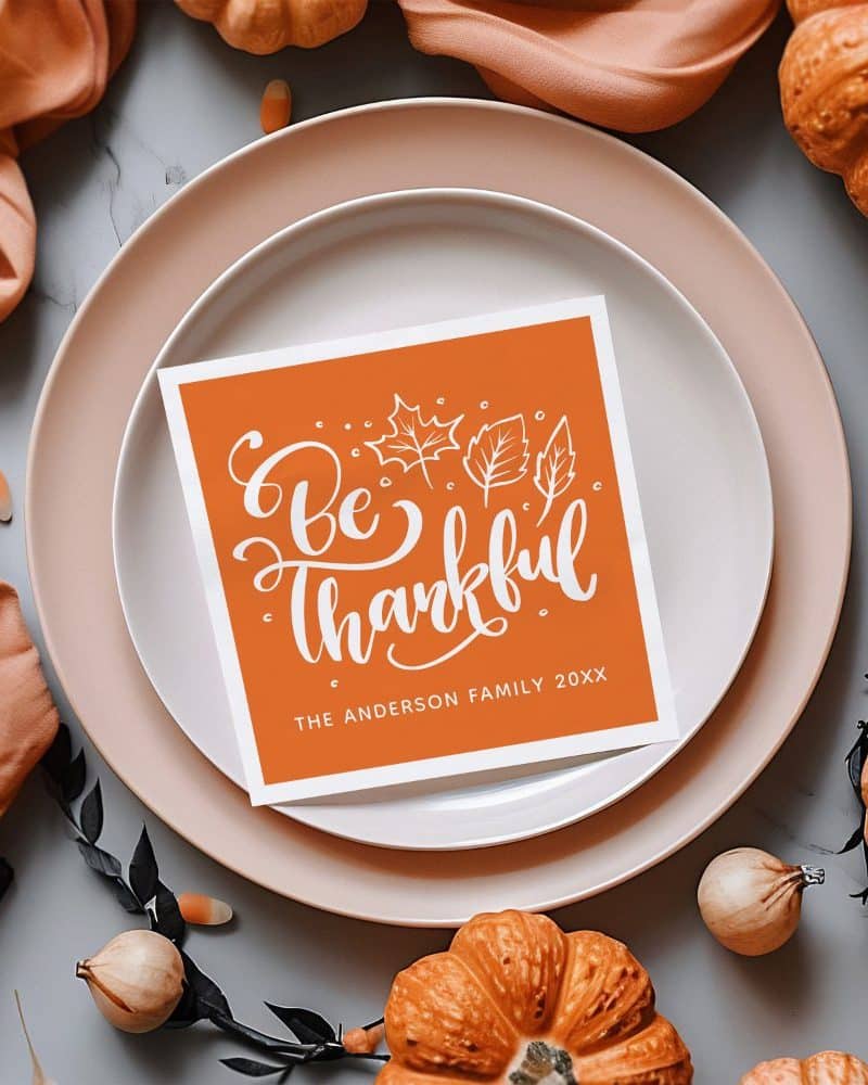 An orange Thanksgiving place card with the words Be Thankful and the Anderson Family name. Small white leaves are depicted around the text.
