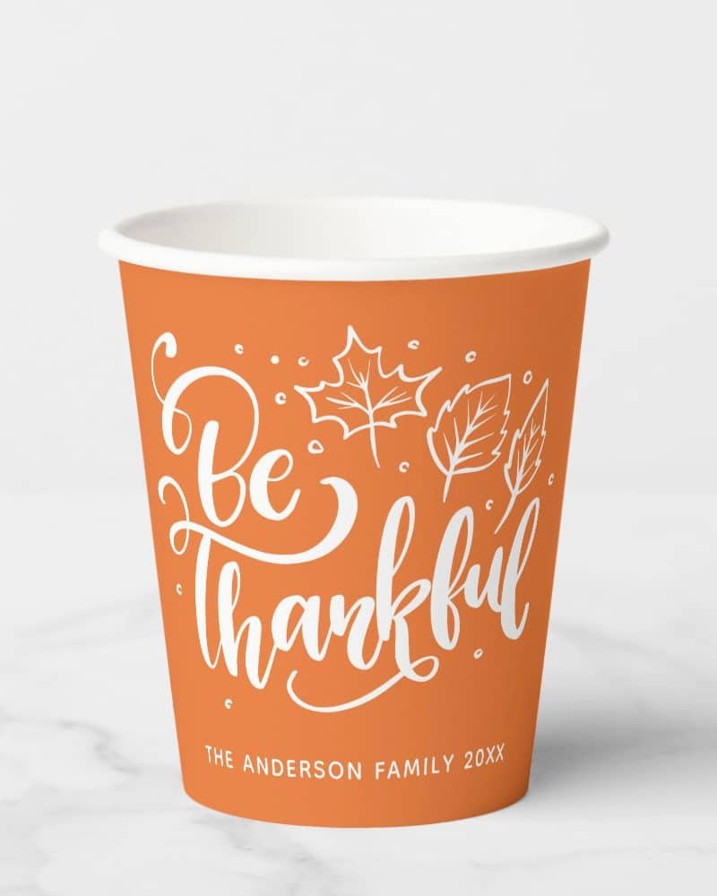 Orange paper cup with white lettering that says Be Thankful and includes a simple leaf design.