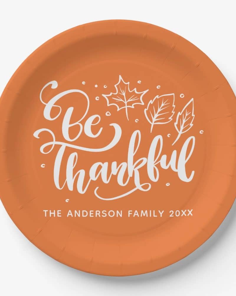 An orange paper plate with the phrase Be Thankful in white script lettering and small white leaf designs. The Anderson Family 20XX is printed below.