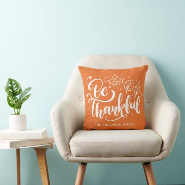 Orange Throw Pillow With White Lettering That Says Be Thankful And The Anderson Family. Leaves Are Also Depicted.