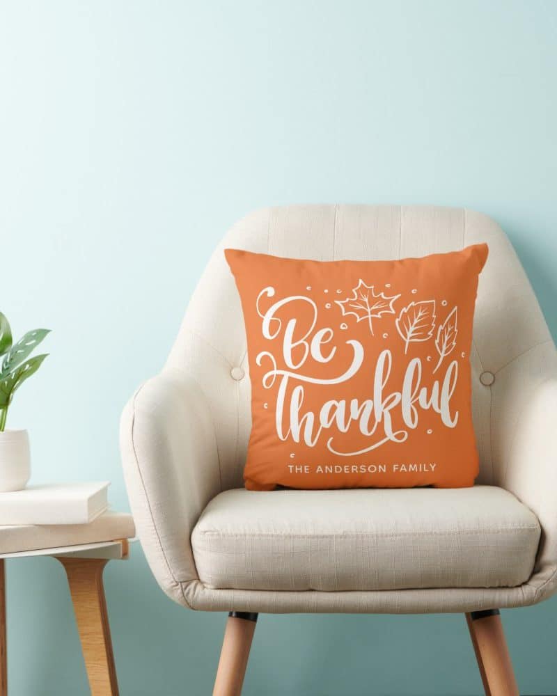 Orange throw pillow with white lettering that says Be Thankful and The Anderson Family. Leaves are also depicted.