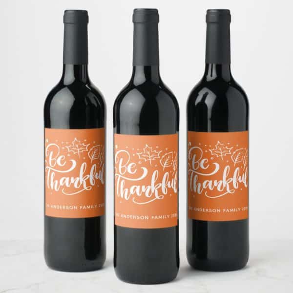 Three Wine Bottles With Orange Labels That Say Be Thankful In A Script Font, With Small Leaf Drawings.