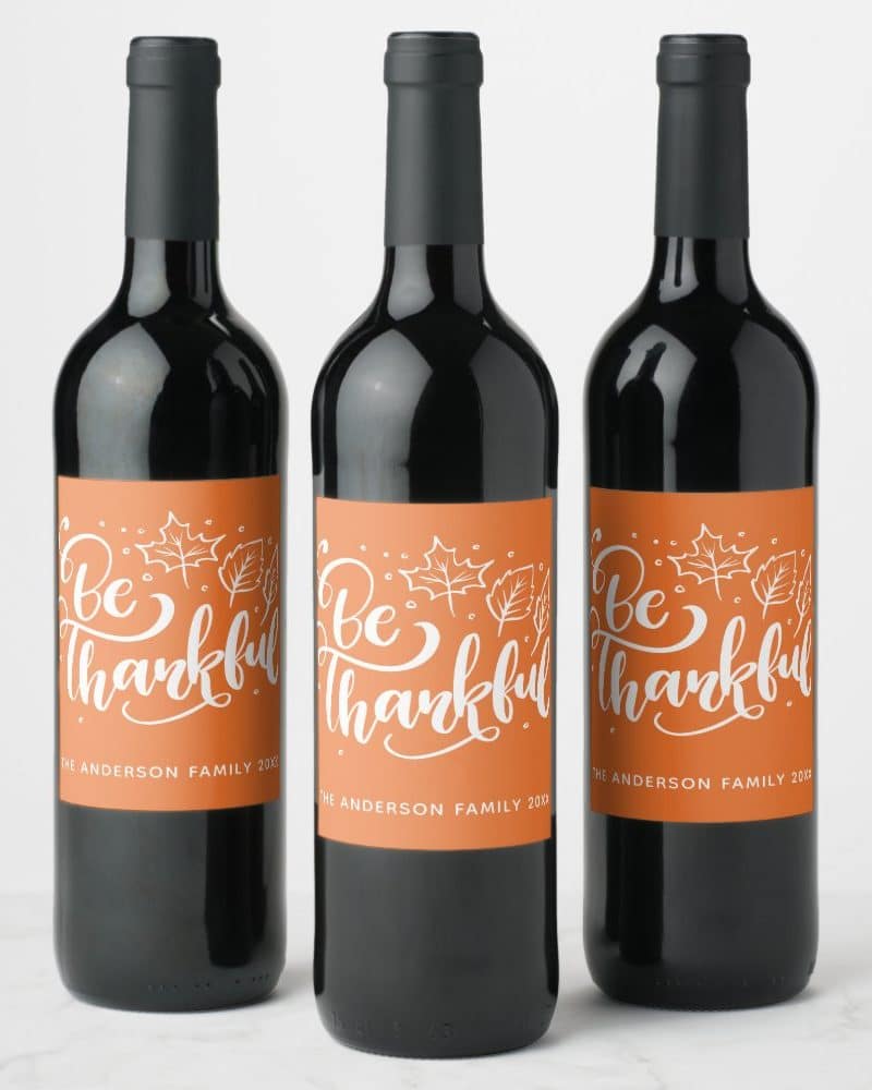 Three wine bottles with orange labels that say Be Thankful in a script font, with small leaf drawings.