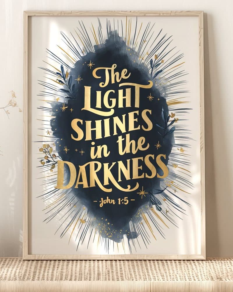 Framed print with gold lettering on a dark blue watercolor background. The text reads The Light Shines in the Darkness, John 1:5.