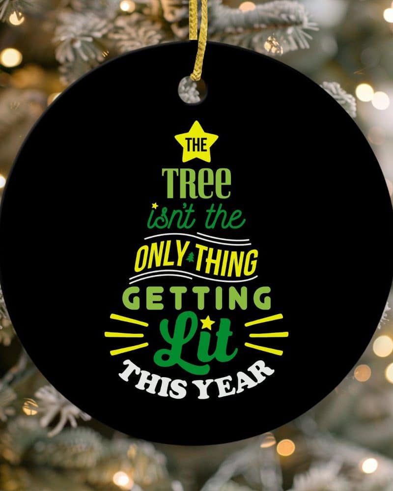 Round black Christmas ornament with green and yellow text that reads, The tree isnt the only thing getting lit this year.