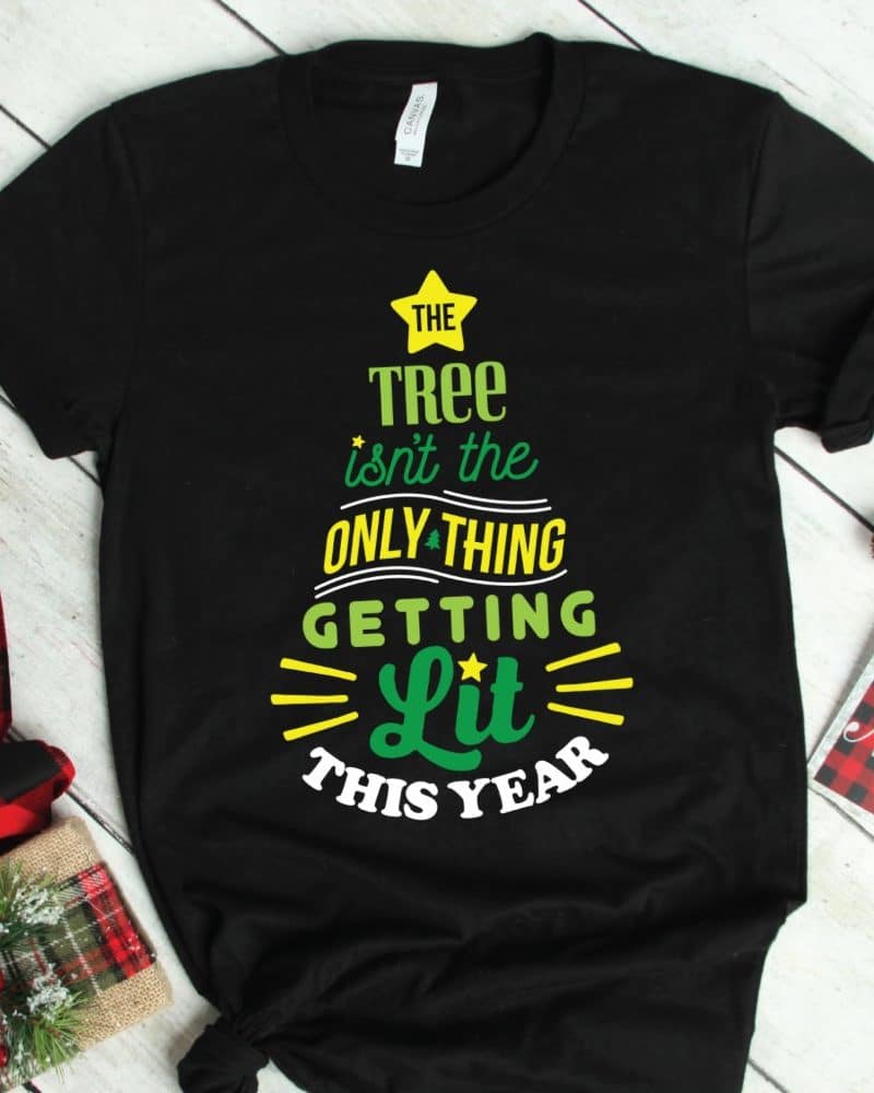 Black t-shirt with a Christmas tree design. The tree is formed by text that says The Tree isnt the Only Thing Getting Lit This Year.