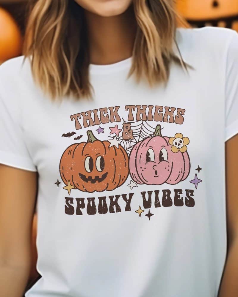 White t-shirt with a Halloween design featuring two cartoon pumpkins and the text Thick Thighs & Spooky Vibes.