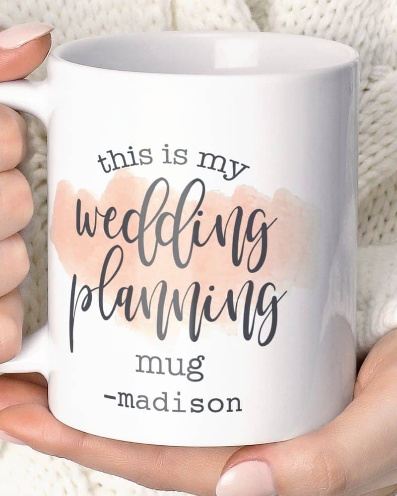 A white coffee mug with the text This is my wedding planning mug -madison on it. A peach watercolor design is behind the words wedding planning.