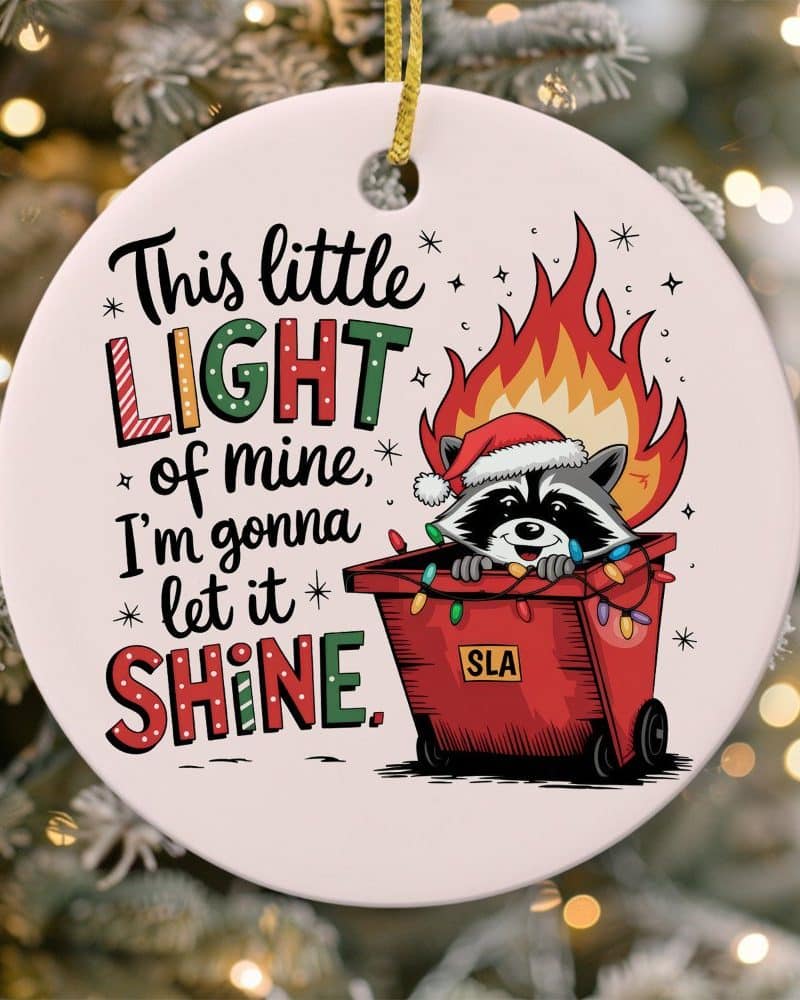 Christmas ornament featuring a raccoon wearing a Santa hat inside a dumpster decorated with Christmas lights and flames, with text reading This little LIGHT of mine, I'm gonna let it SHINE.
