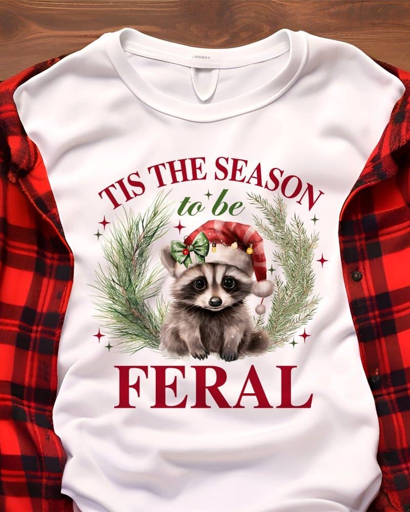 White sweatshirt with a Christmas raccoon wearing a Santa hat in a wreath design. The text reads Tis the Season to be Feral.