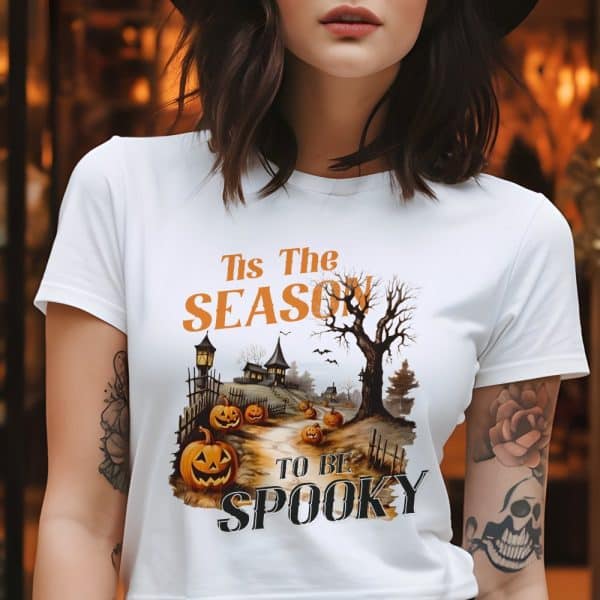 Woman Wearing A White T-Shirt With A Halloween Design. The Design Features A Spooky Village Scene With Jack-O'-Lanterns And The Text Tis The Season To Be Spooky.