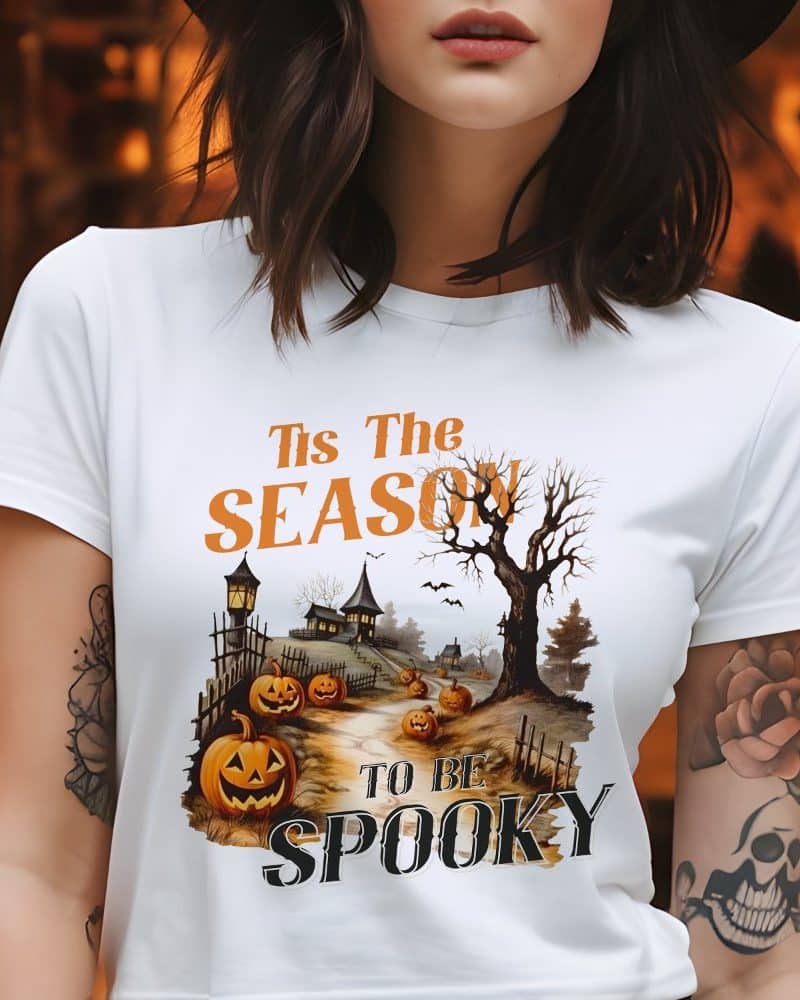 Woman wearing a white t-shirt with a Halloween design. The design features a spooky village scene with jack-o'-lanterns and the text Tis the Season to be Spooky.
