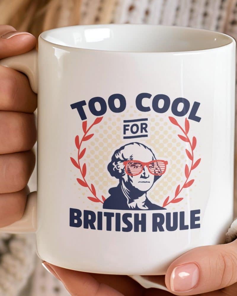 White coffee mug with a graphic of George Washington wearing red sunglasses, surrounded by a laurel wreath; text reads "Too Cool For British Rule".