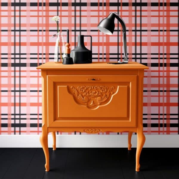 Orange Painted Wood Cabinet With Ornate Detailing, Holding A Lamp And Decorative Items, Sits Against A Pink And Black Plaid Wall.
