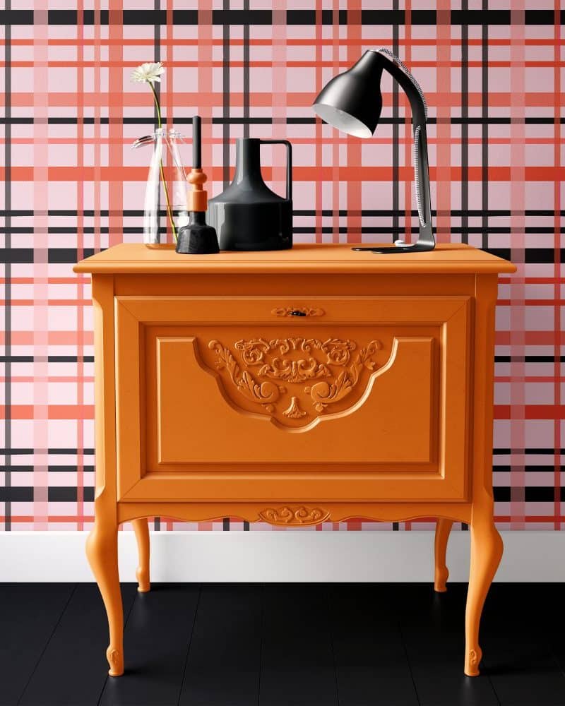 Orange painted wood cabinet with ornate detailing, holding a lamp and decorative items, sits against a pink and black plaid wall.